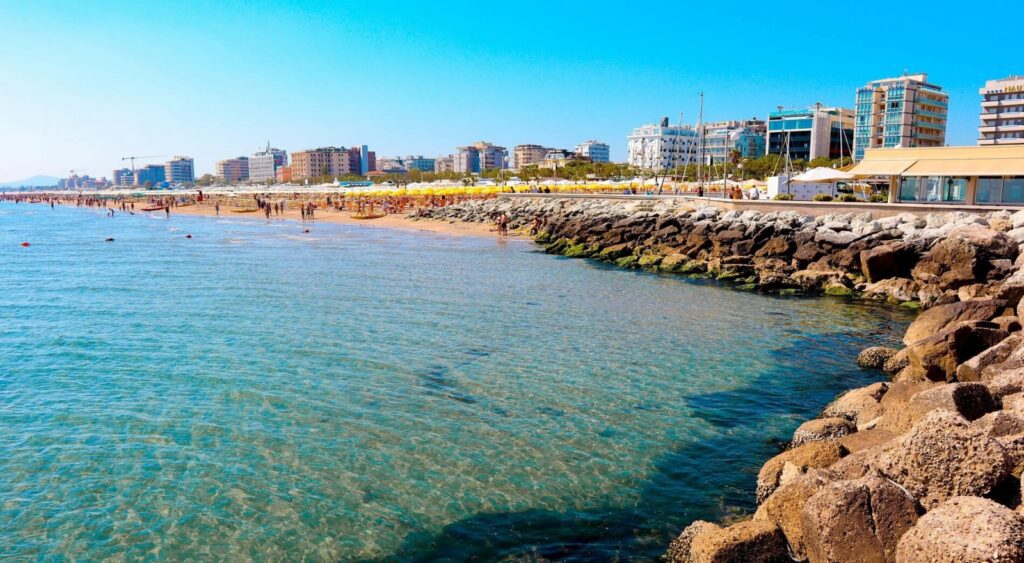 Spiaggia affollata con edifici sul lungomare e mare limpido.
